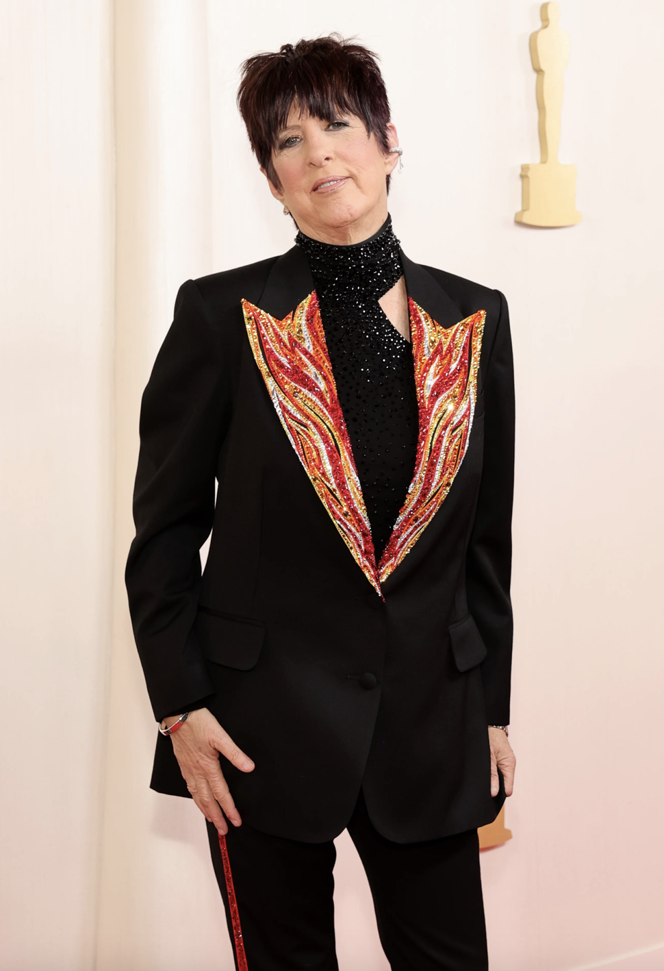 A woman with short dark hair stands wearing a black shirt and suit with red detailing on the lapels.