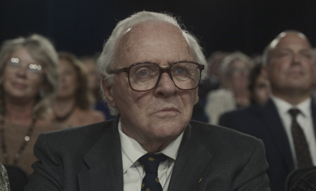 An elderly man with a receding white hairline sits in the front row of an audience wearing a suit and spectacles.