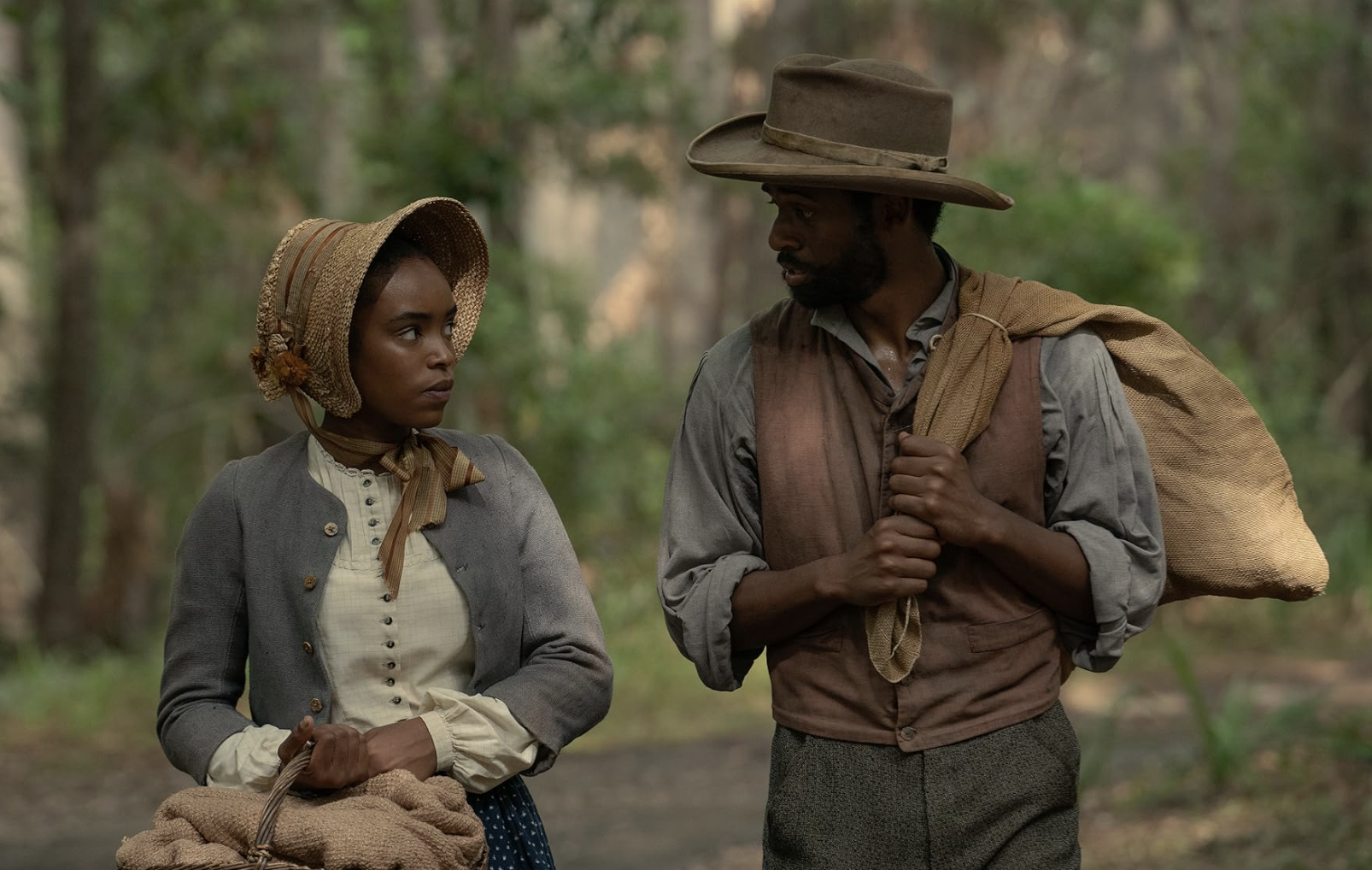 A Black woman wearing a bonnet and carrying a basket walks through the woods with a Black man who is carrying a sack and wearing period clothing from the 1860s.