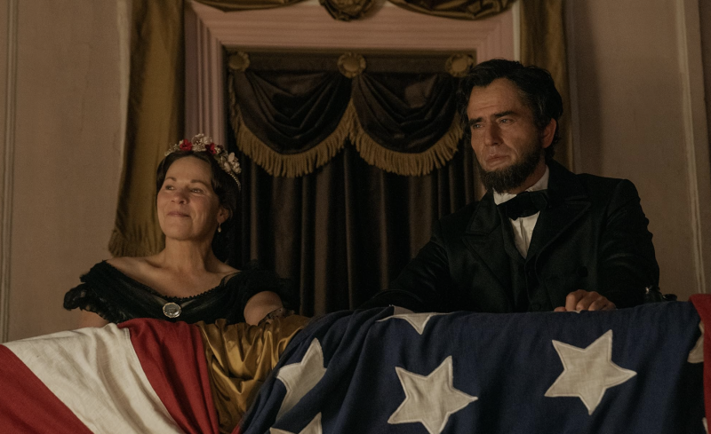 Actors playing President Lincoln and his wife sit in a theater box, an American flag draped in front of them.