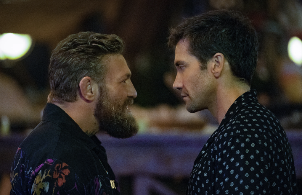 A square-jawed white man stares into the face of a shorter, bearded white man, as if they are about to fight.