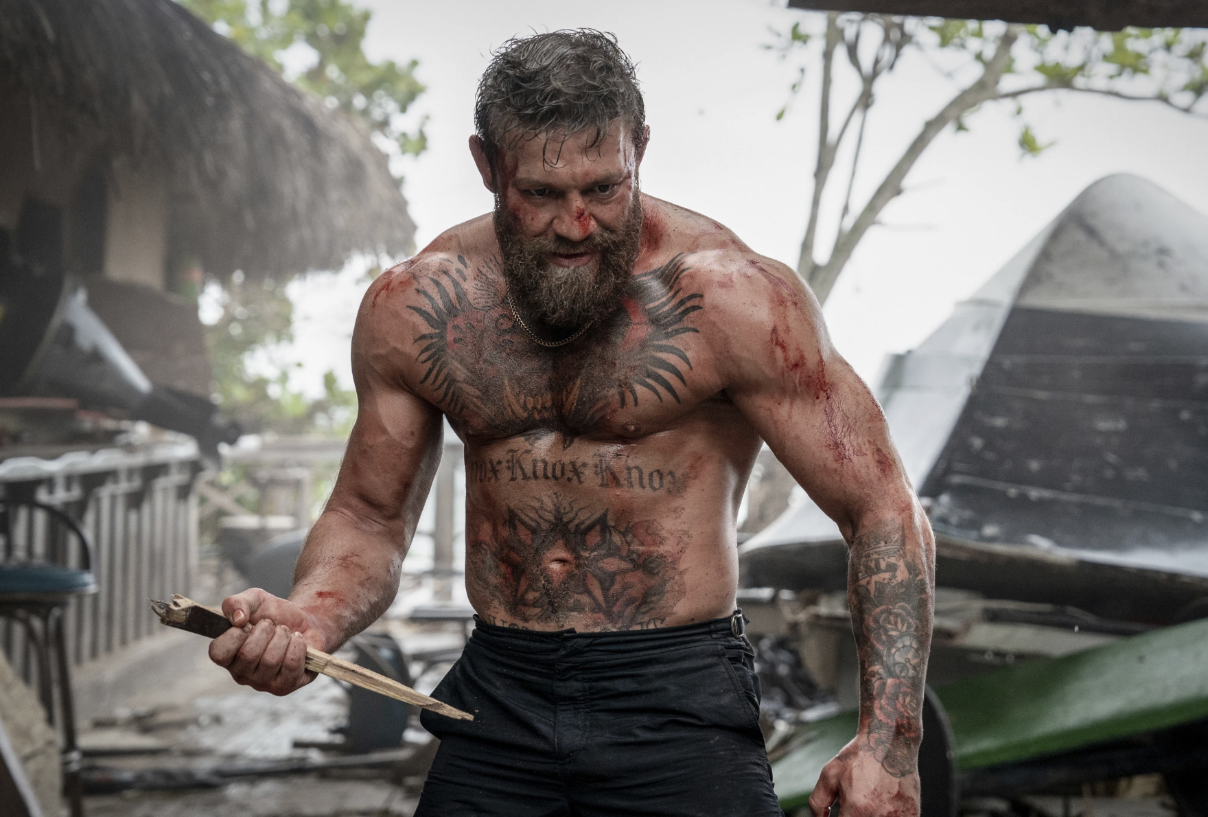 A shirtless and very muscular bearded man stands poised to fight, clutching a dagger.