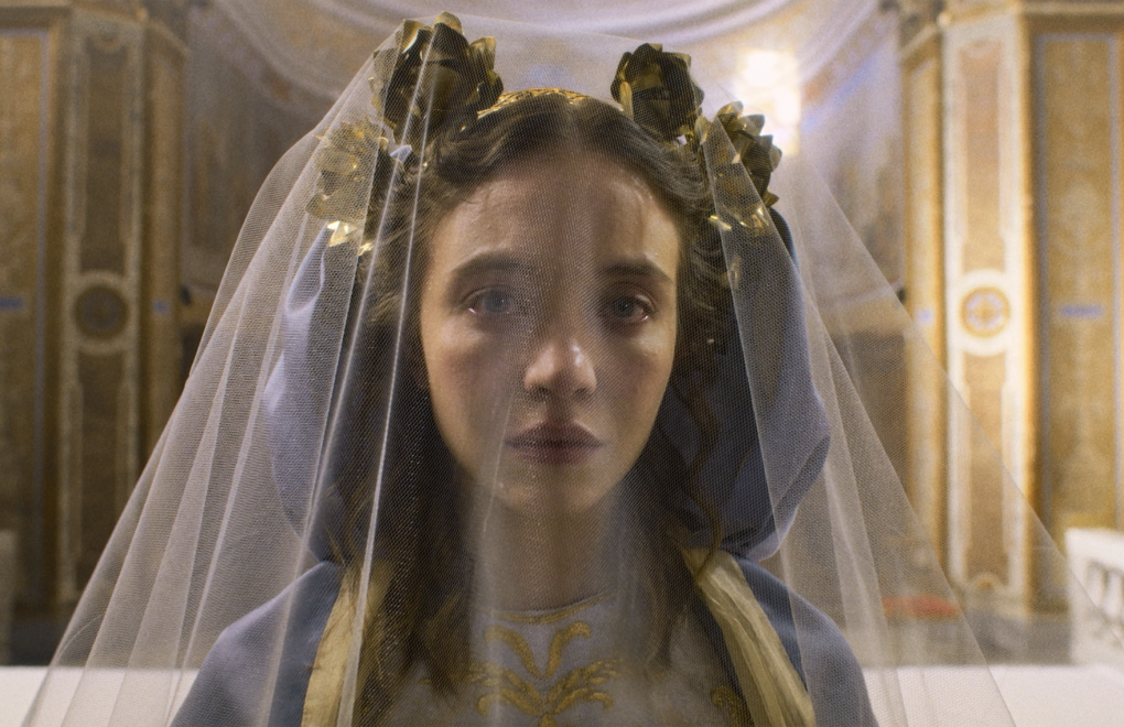 A white woman with gold hair accessories looks sullen from beneath a thin white veil.