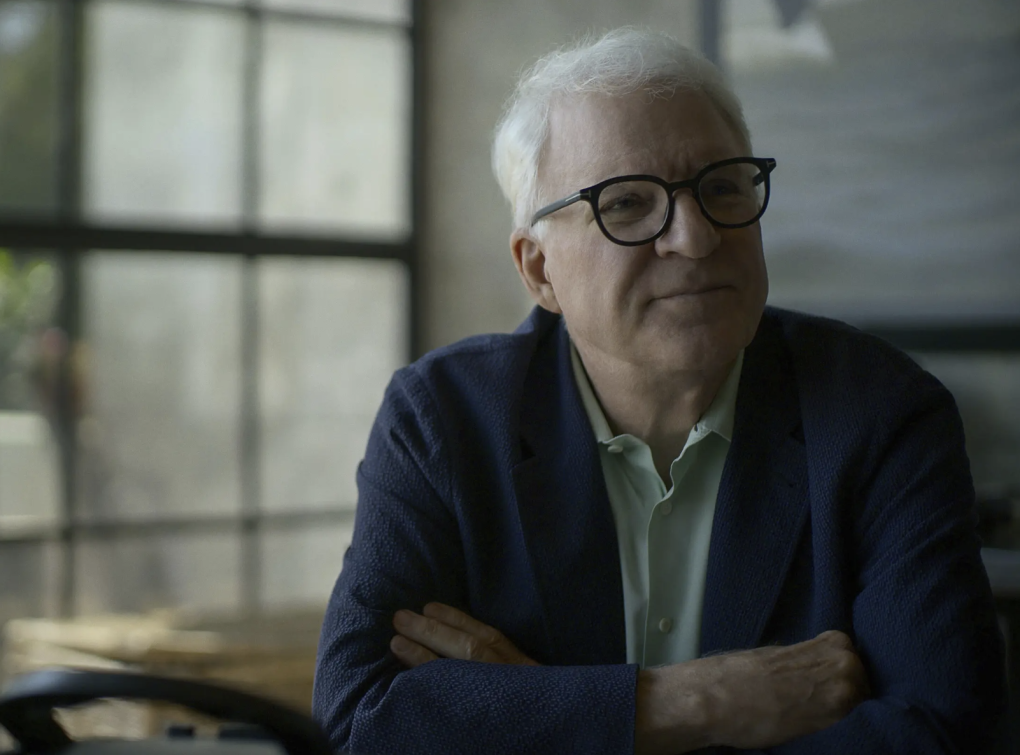 A white-haired man in glasses sits at a desk with his arms folded. A bright window lines the wall behind him.