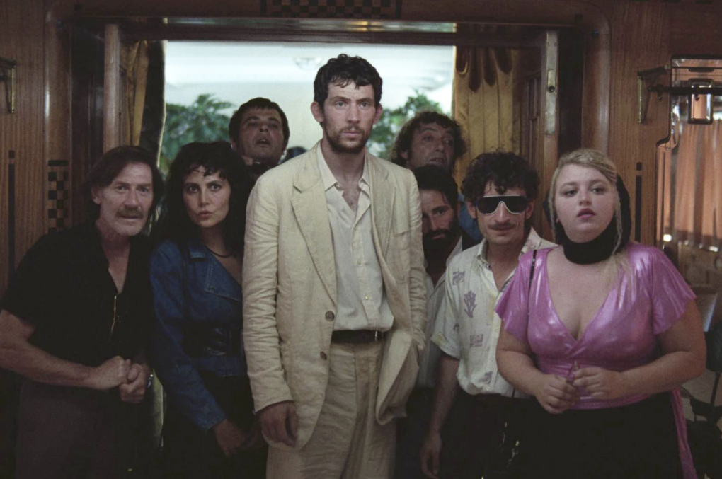 A white man in a beige linen suit stands in a hallway surrounded by a mix of oddball men and women.
