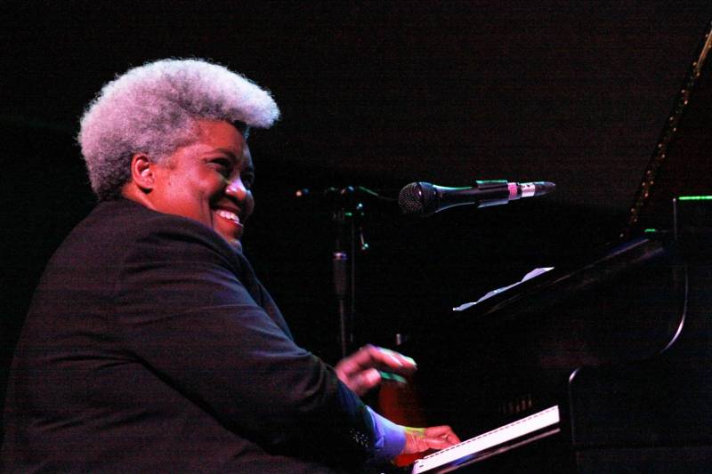 A Black woman in black top and greyish hair styled up plays the piano.