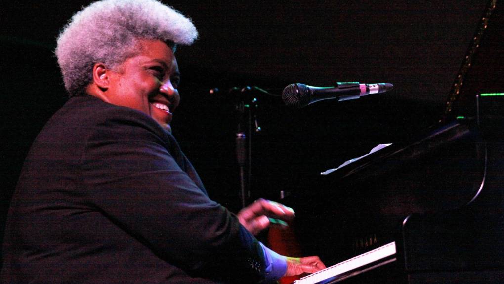 A Black woman in black top and greyish hair styled up plays the piano.