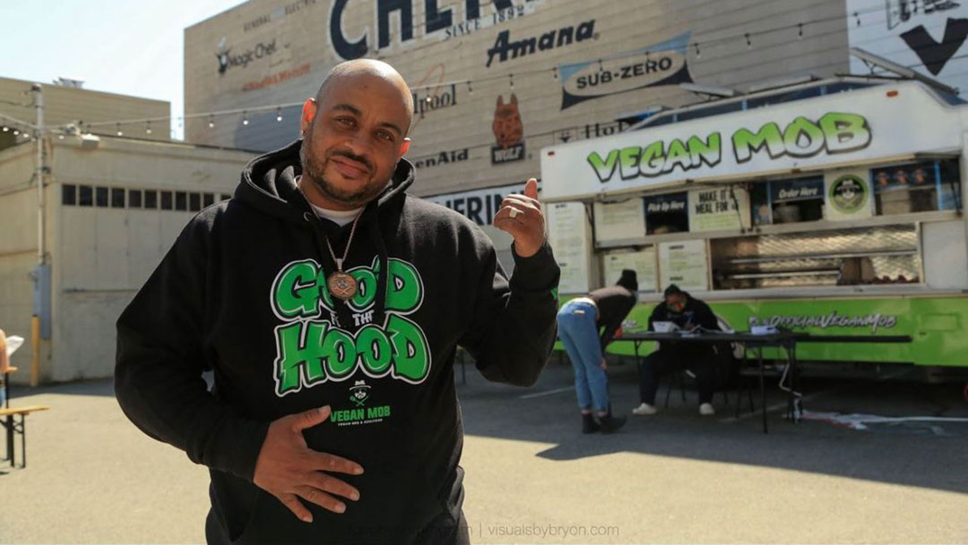 Man in a black "Good Hood" sweatshirt gestures toward the Vegan Mob food truck behind him.