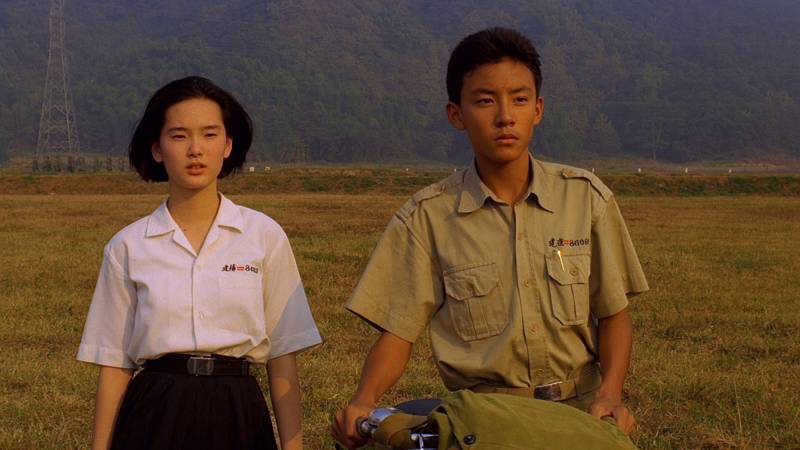 Still shot from a movie: A teenage boy and girl in their school uniforms walk in an empty field.