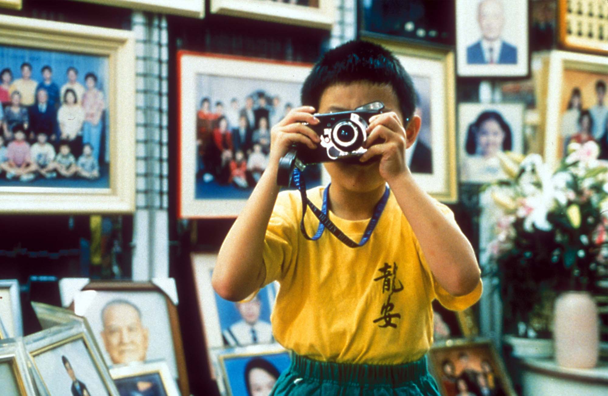 A boy in a yellow T-shirt holds a camera to his face.