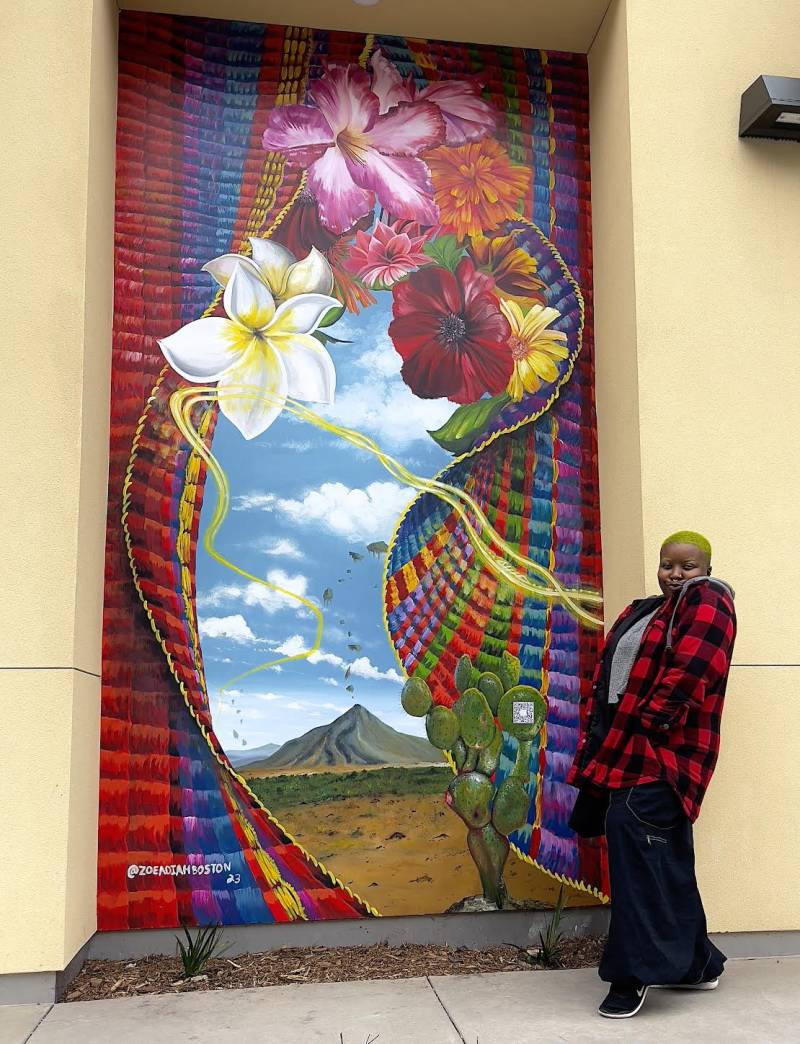 Zoë Boston wears a plaid long-sleeve shirt as she poses for a photo in front of her mural on the Casa Sueños Apartments in East Oakland.