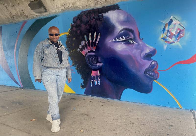 Zoë Boston wears sunglasses and a denim outfit as she stands in front of a mural depicting a beautiful purple being on a blue background.
