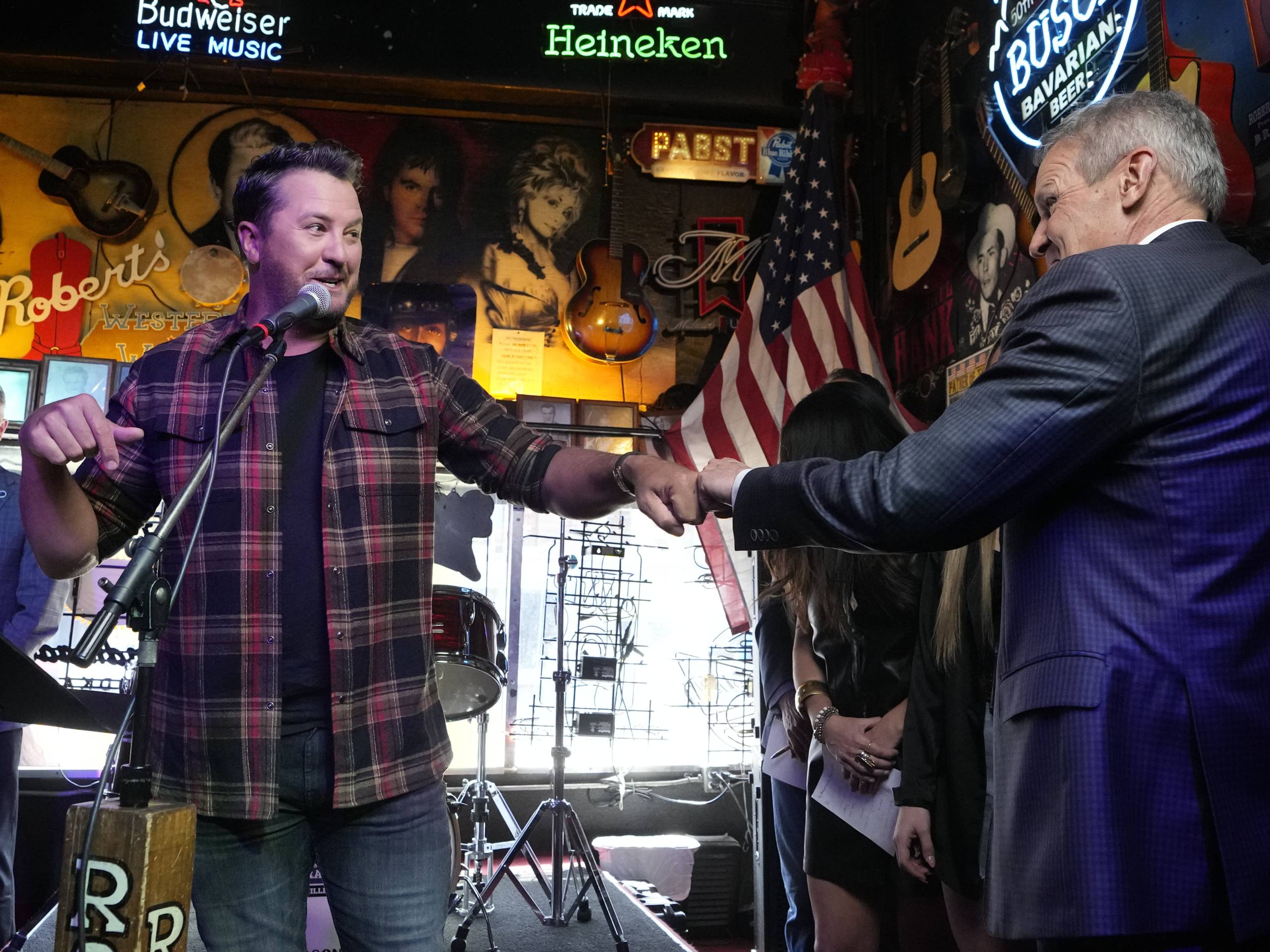 A man in a flannel patterned shirt gives a fist-bump to a man in a suit as the American flag hangs in the background, indoors
