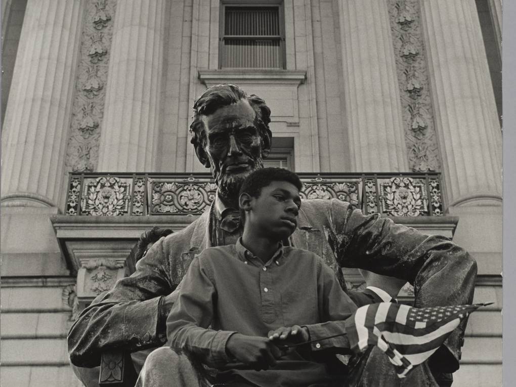 Black-and-white photo of young person sitting with American flag in front of Lincoln statue
