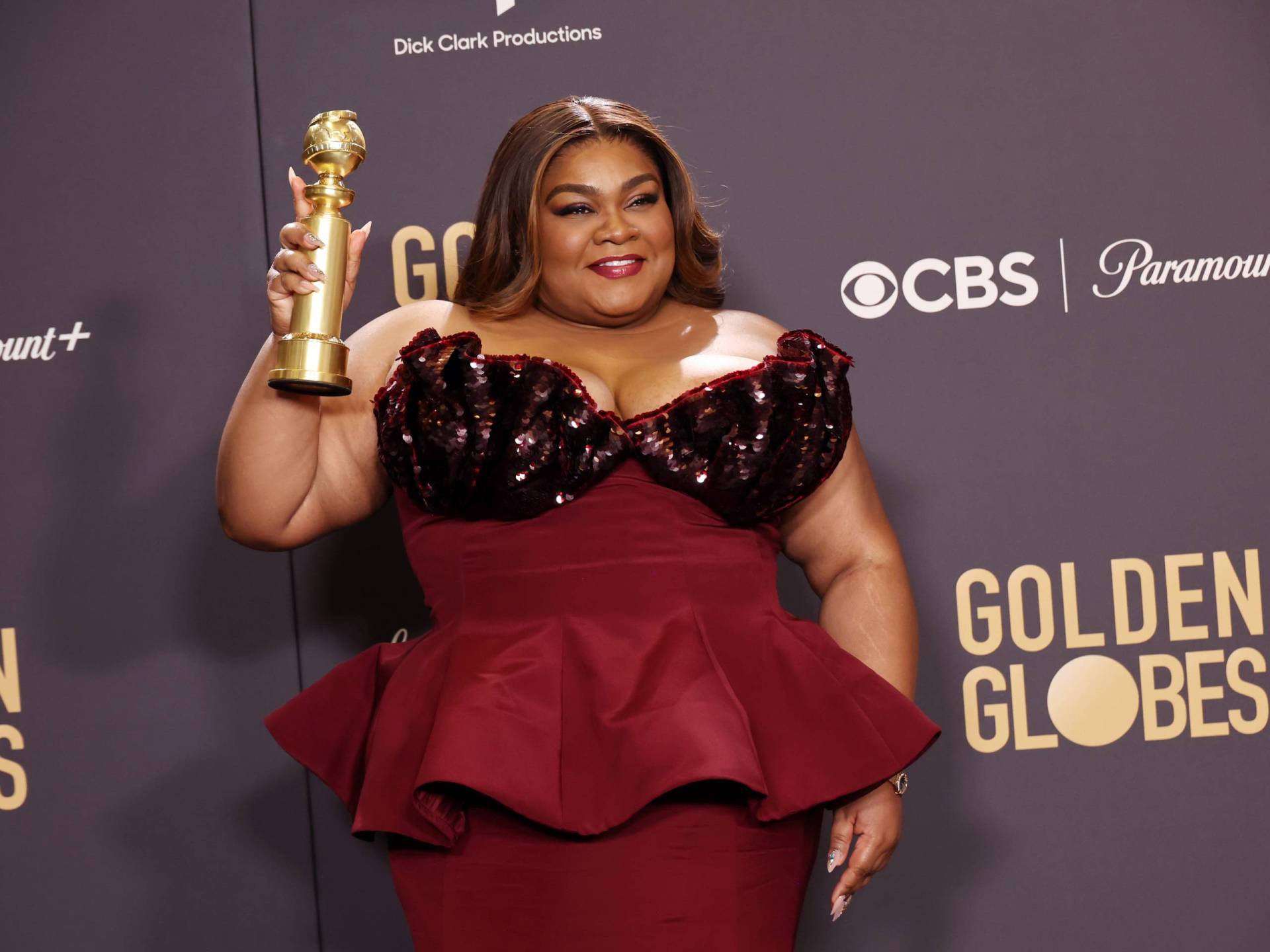 A plus-sized Black woman in a red gown holds up a trophy.