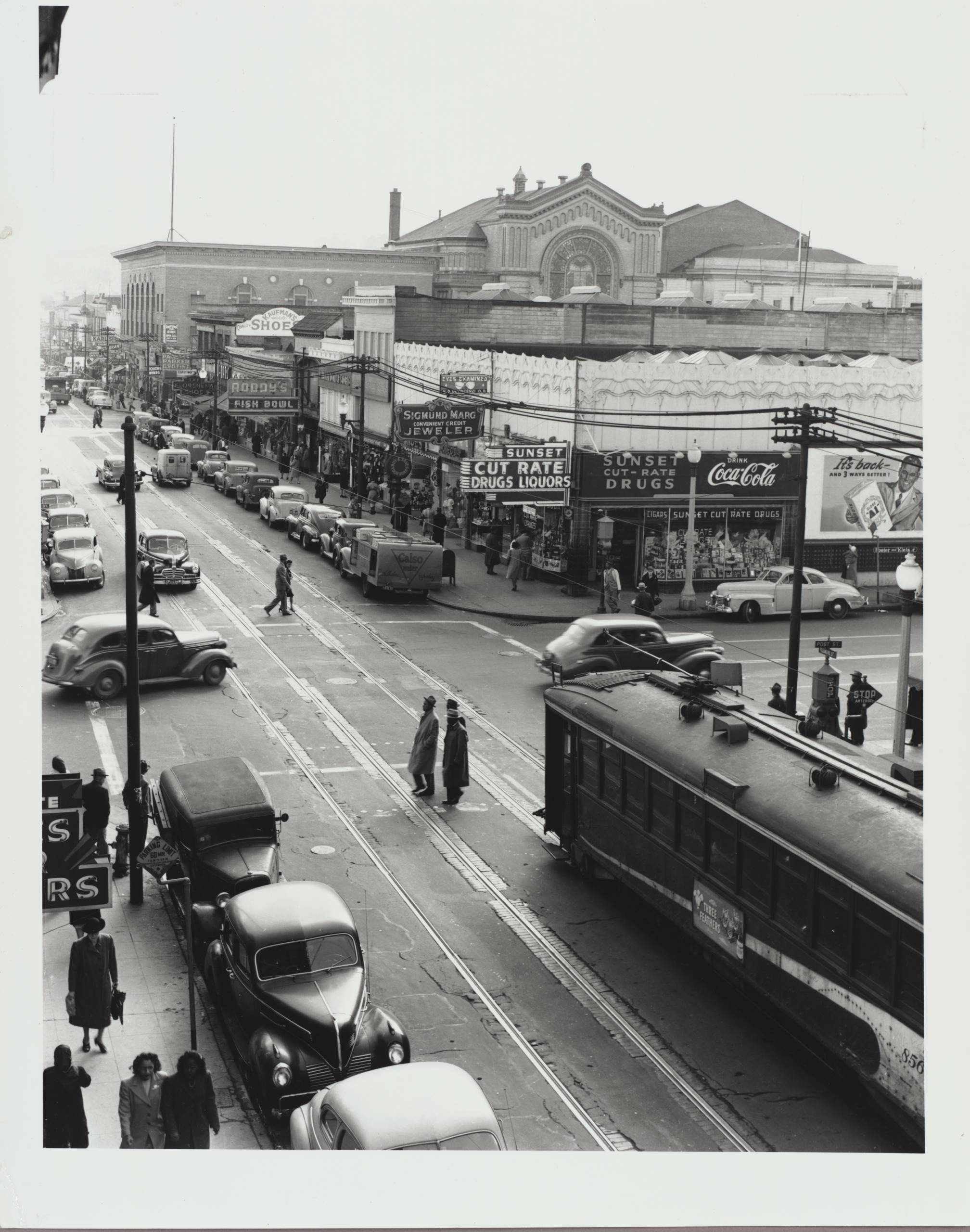 Black-and-white photograph of busy street corner with pedestrian, car and bus traffic