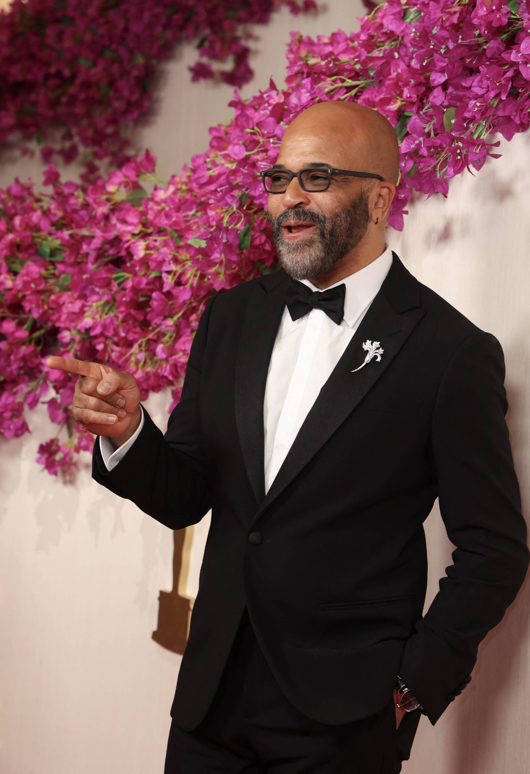 A bald Black man wearing glasses and a neat grey beard gestures on a red carpet. He's wearing a tuxedo.