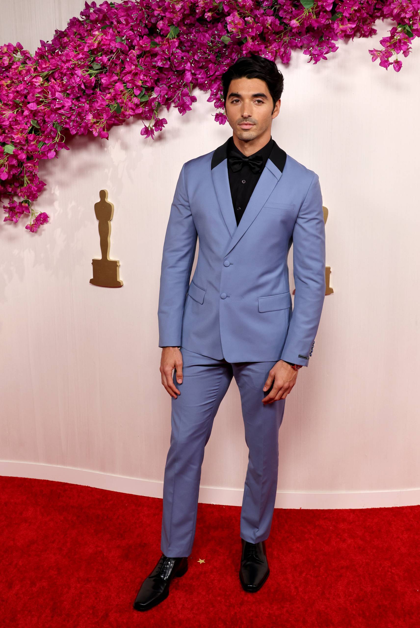 A slight man stands on a red carpet wearing black shirt and cornflower blue suit.