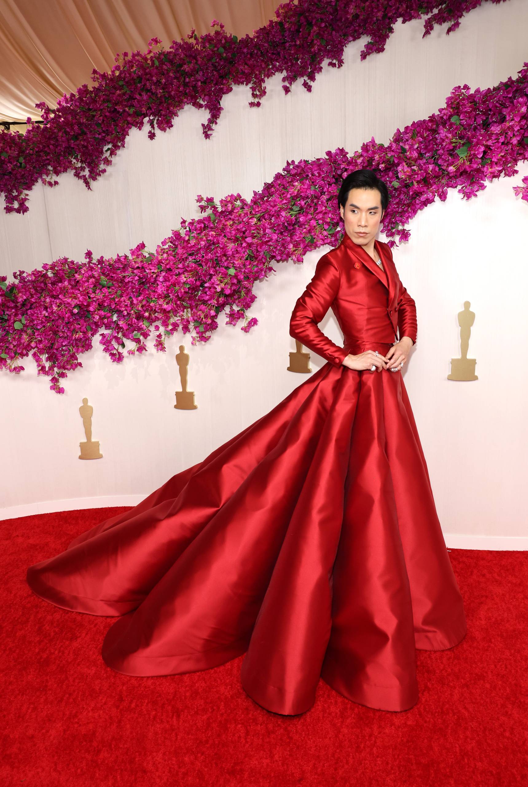 A striking Asian person with dark hair poses, hands on hips, in a red gown with very large skirts.
