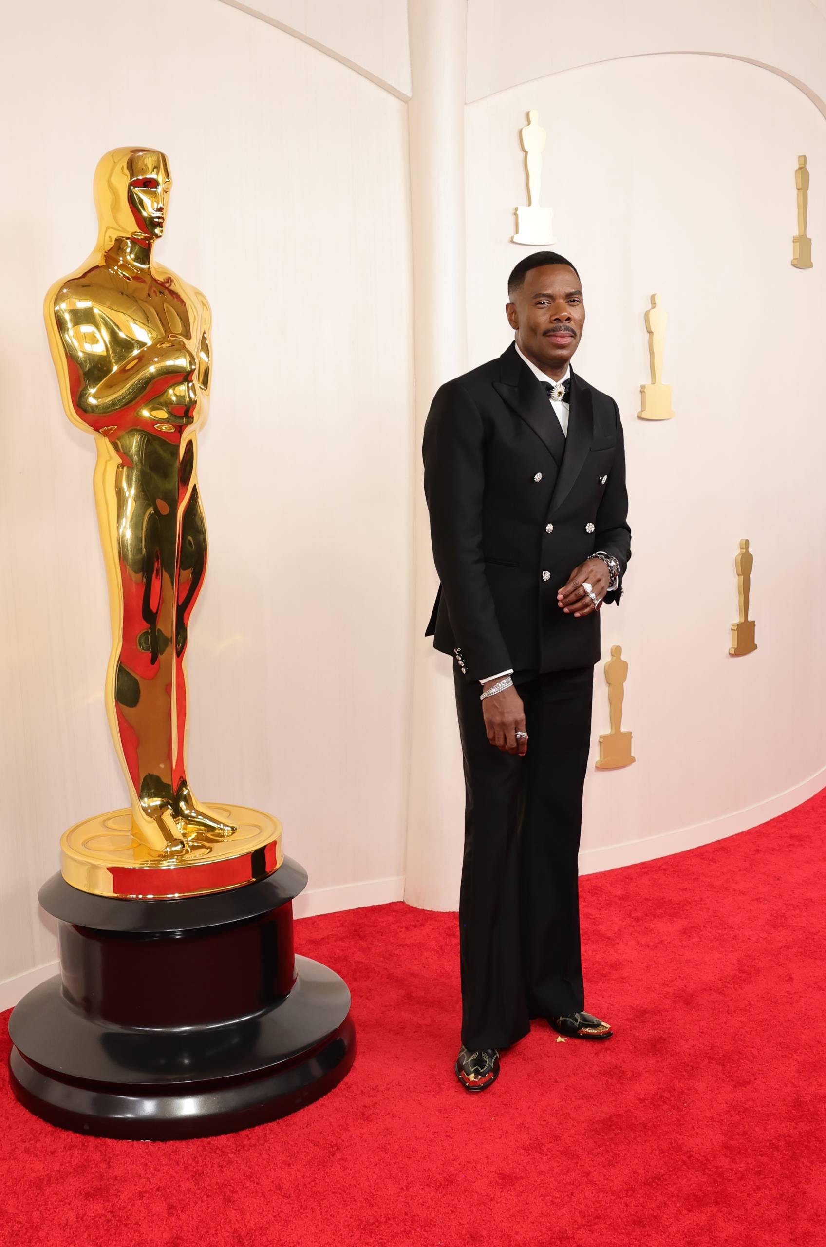 A black man in a black suit and white tie poses next to a life size Oscar statue.