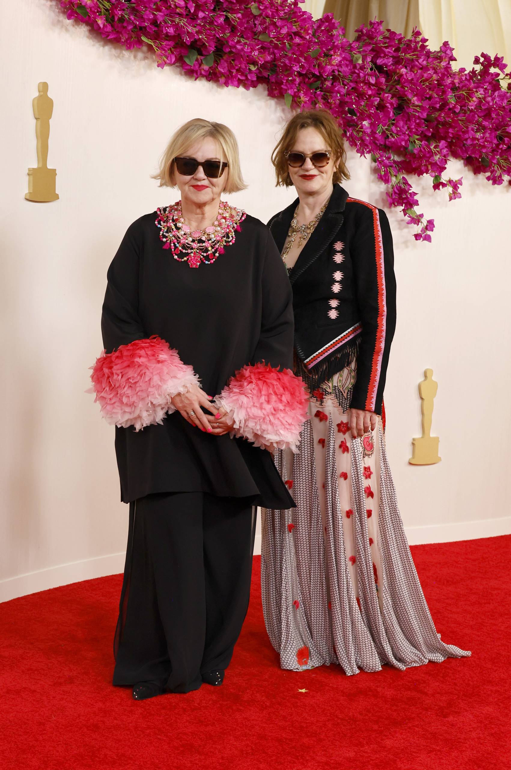 Two white women stand on a red carpet wearing sunglasses. One wears a Black suit with exaggerated pink cuffs. The other wears long floral skirt and military jacket.
