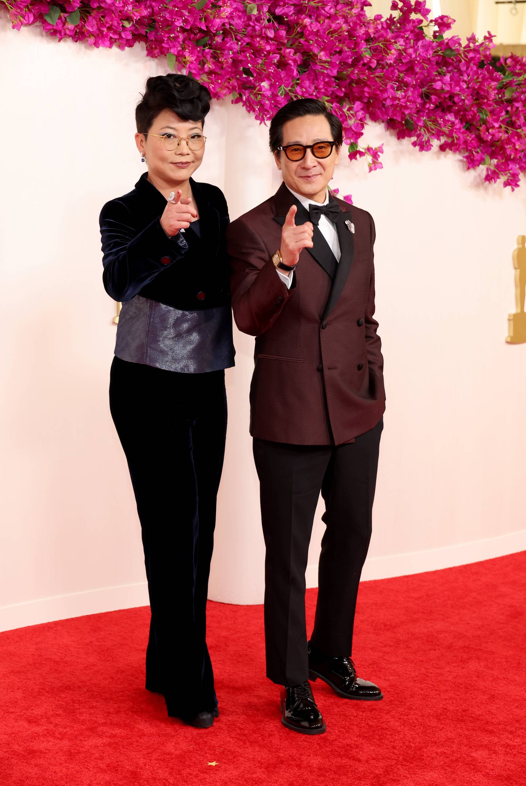 An Asian woman in a black suit stands with an Asian man in a bergundy tuxedo. They are both gesturing to the camera.