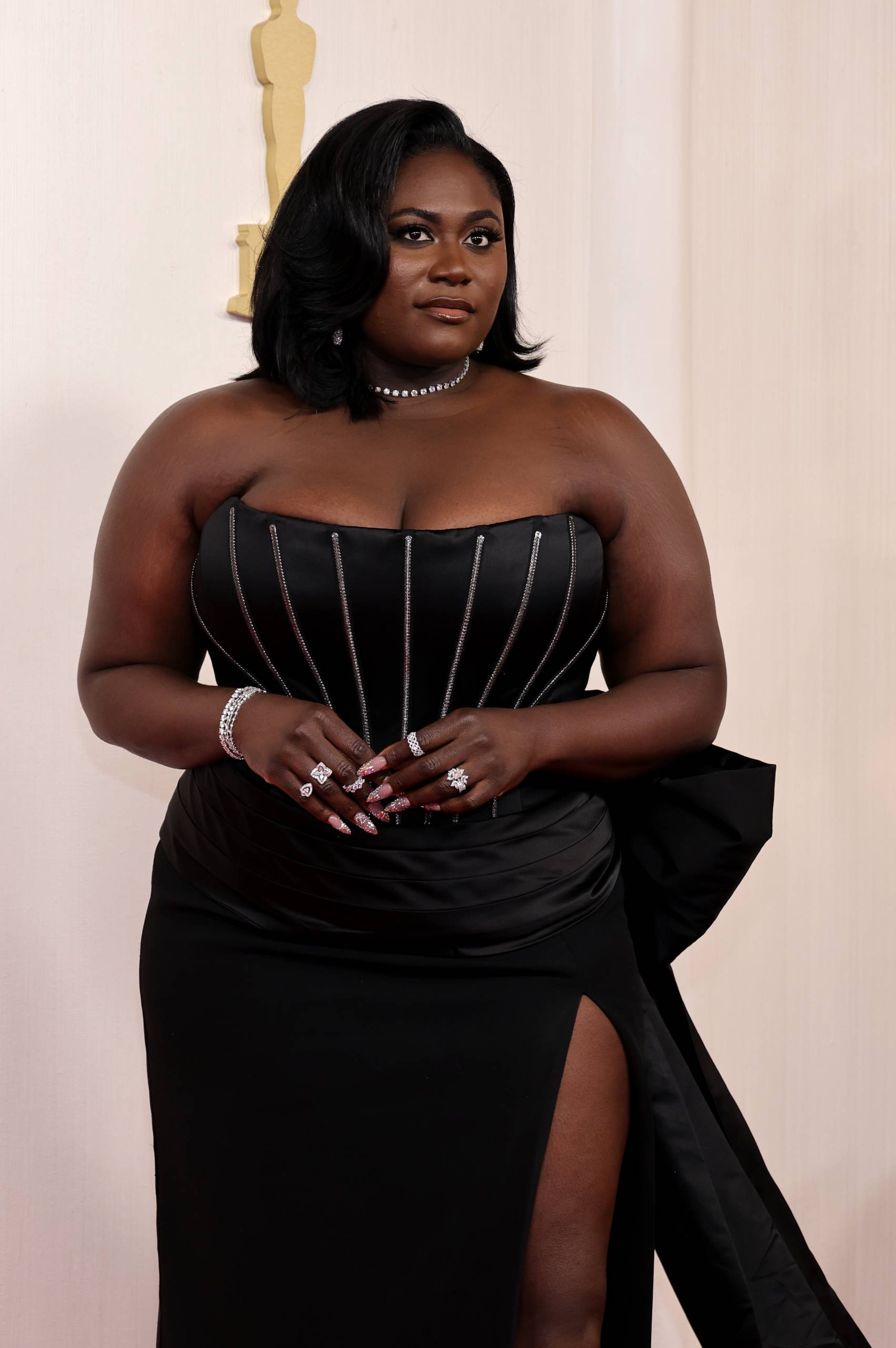 A curvy Black woman stands wearing a strapless black gown with high leg slit.