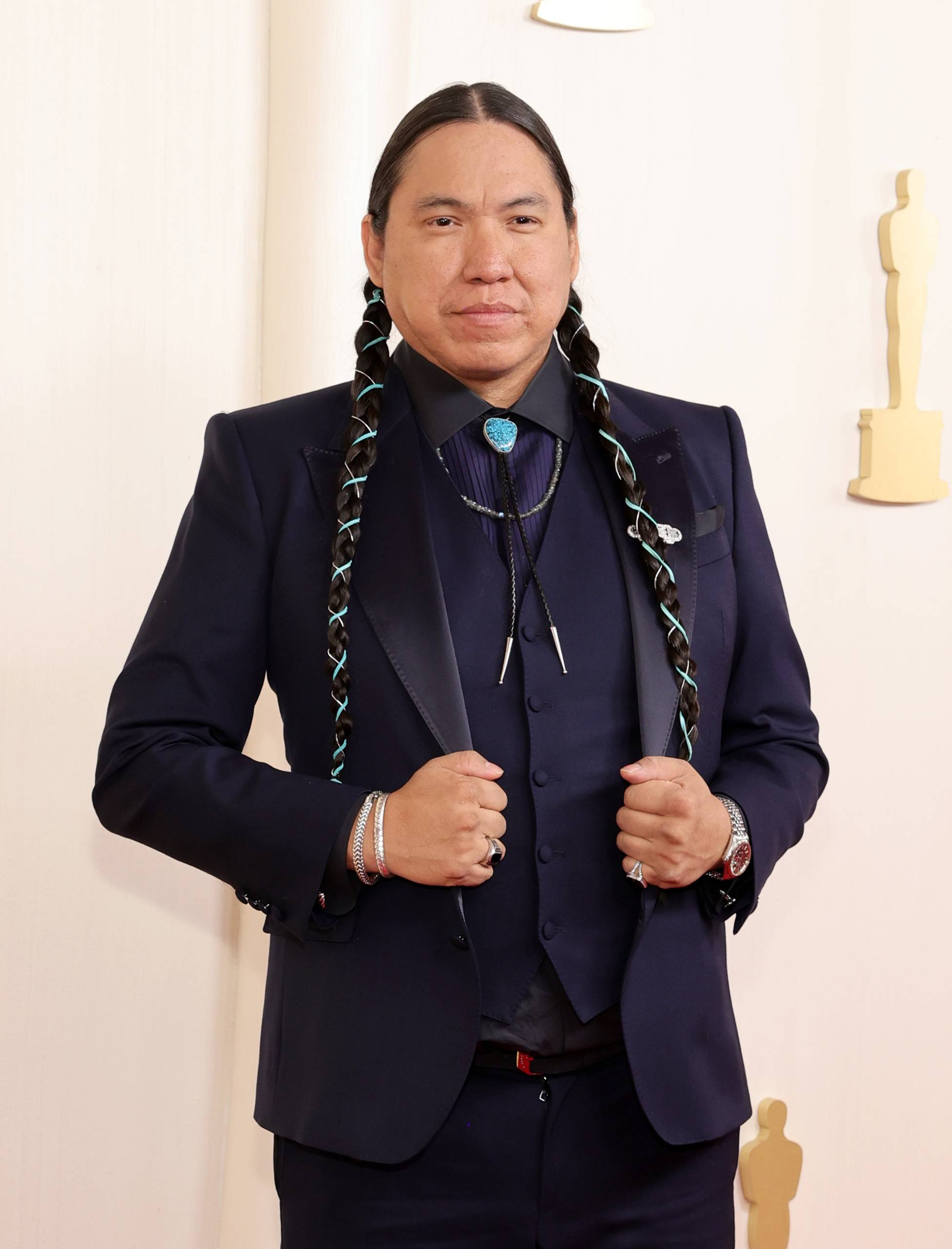 An Indigenous man with long braided hair stands poised wearing a black suit and shirt and a turquoise bolo tie.