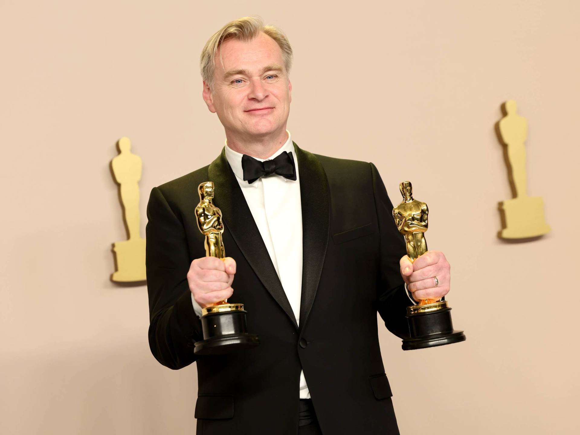 A white man in a tuxedo holds up two gold Oscar statues.