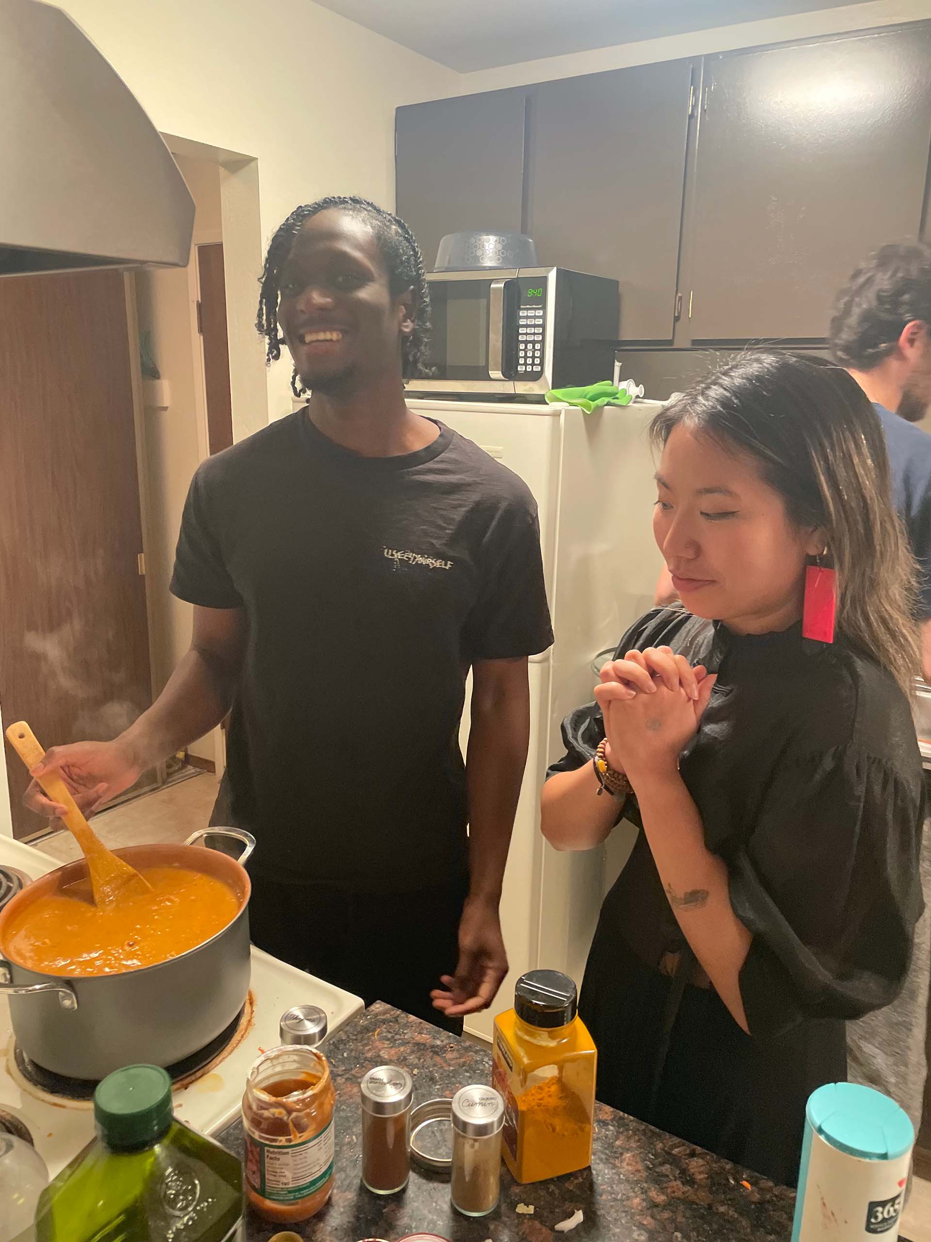 A man stirs a pot of peanut stew while a young woman looks on, pressing her hands together in anticipation.