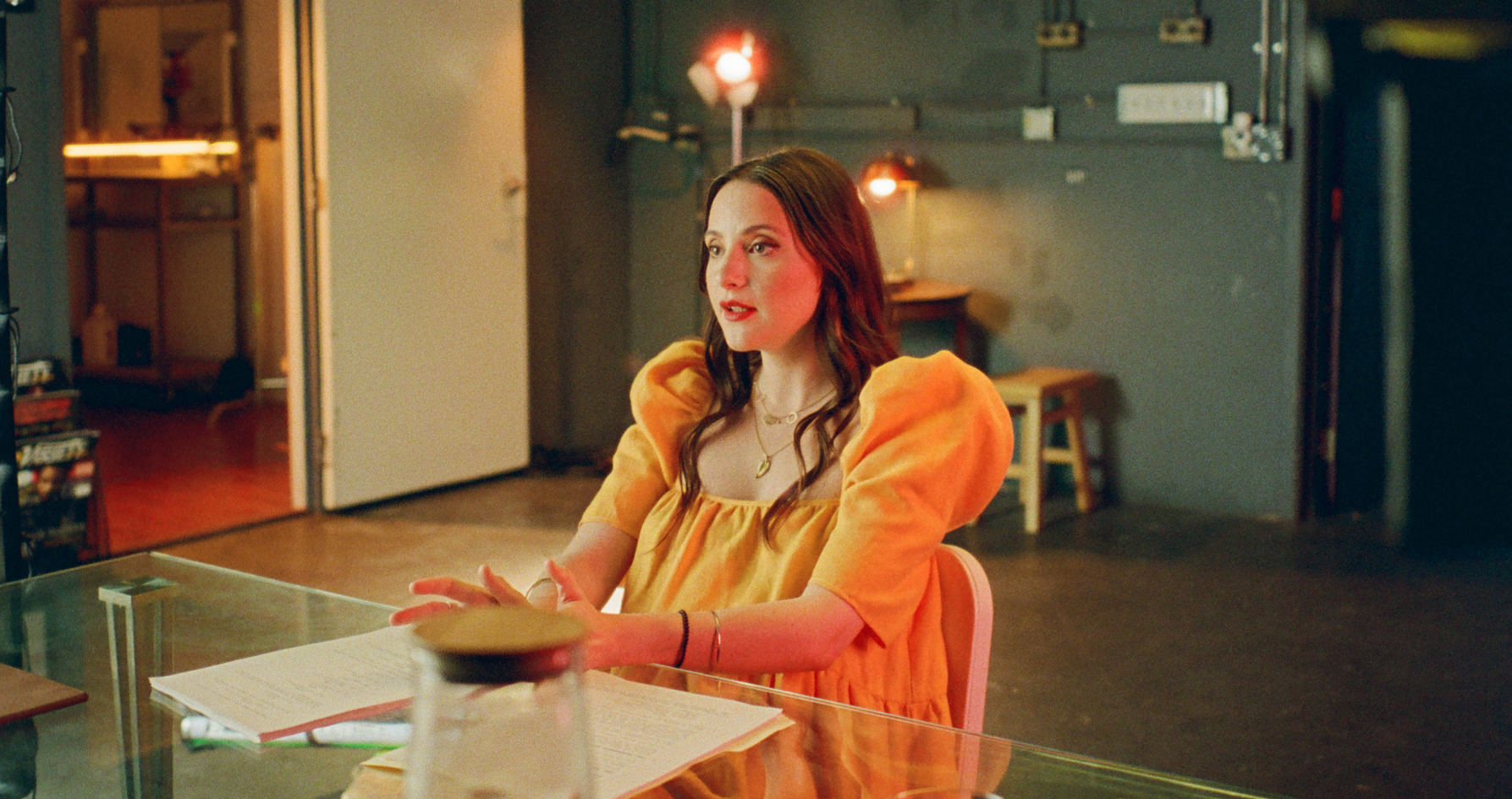 A white woman with long brown hair sits at a transparent table, wearing a striking orange dress with puffy sleeves.