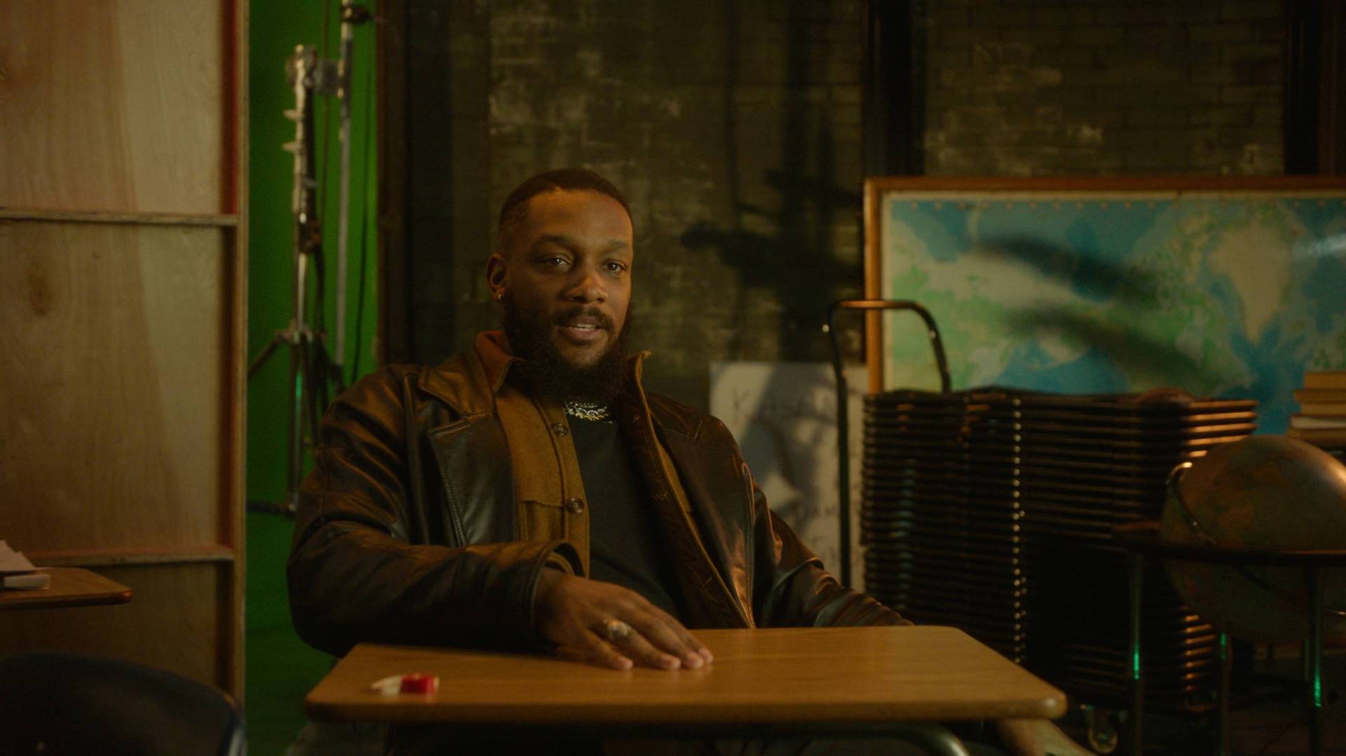 A Black man with a neat beard sits at a school desk in a backstage setting. He is wearing a brown leather jacket and sweater.