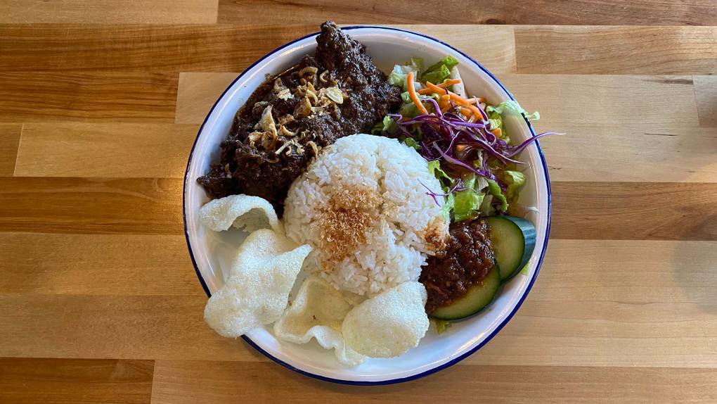 A bowl loaded with beef rendang, coconut white rice, shrimp chips and a salad.