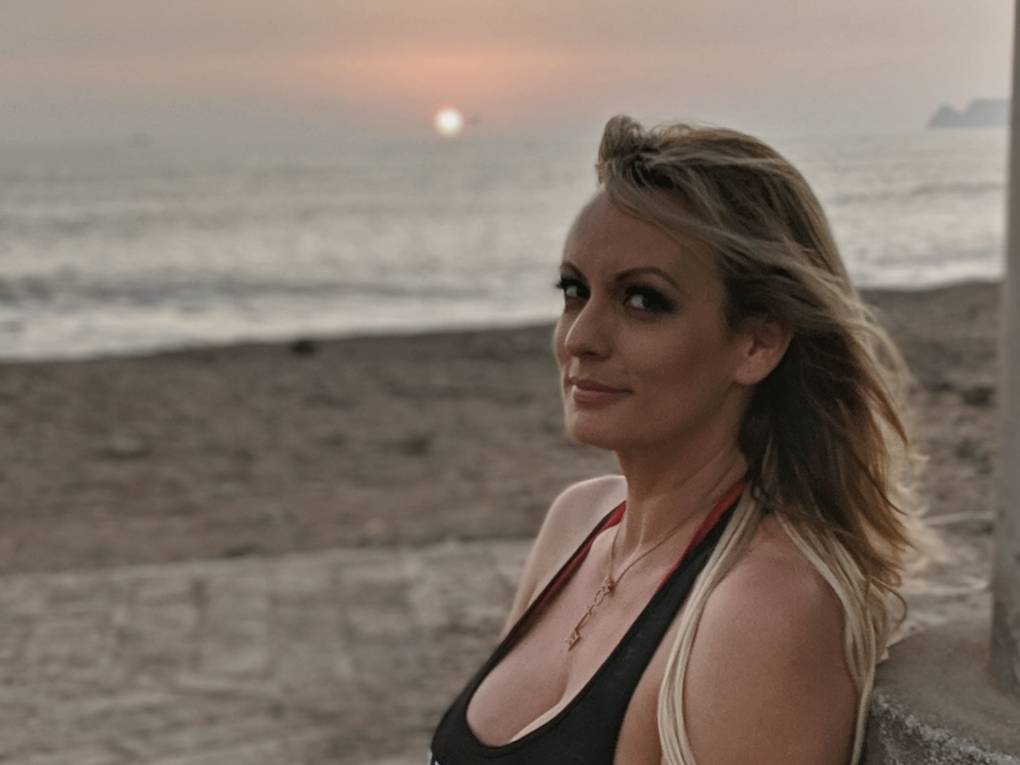 A blonde white woman glances at the camera in side profile, while standing on a beach at sunset.