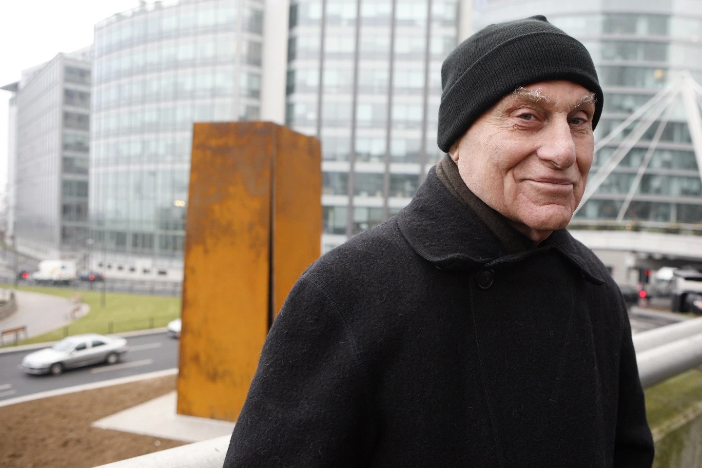 A man in a black jacket and black cap smiles at the camera in front of a bronze/brown sculpture