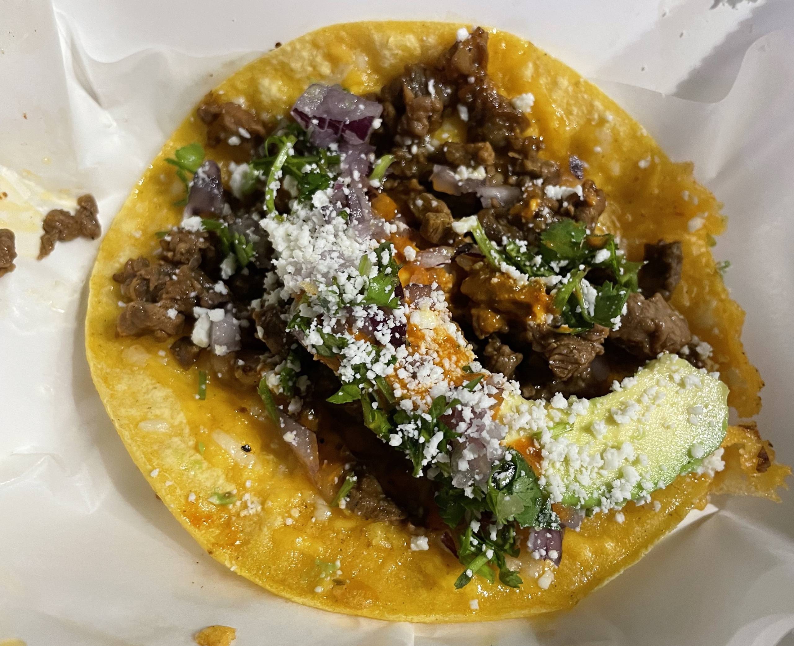 a carne asada taco with guacamole, cilantro and onions on a paper tray