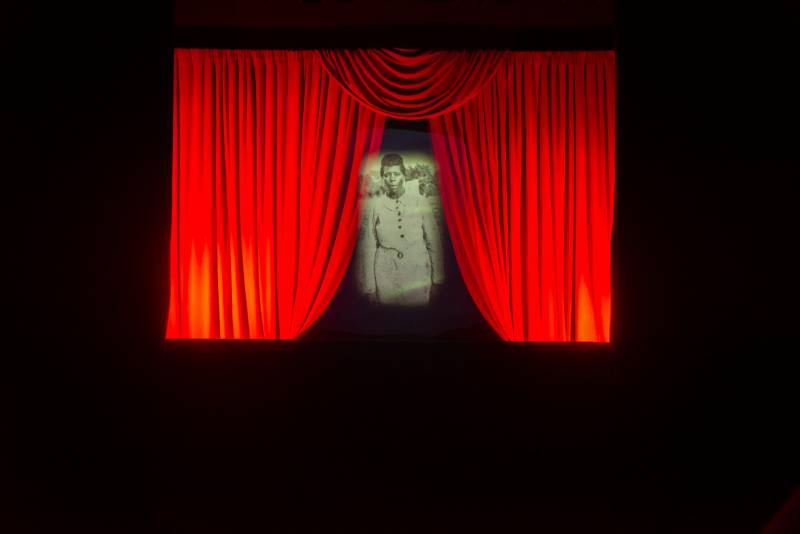 A black and white digital image floats in front of a slightly open red curtain