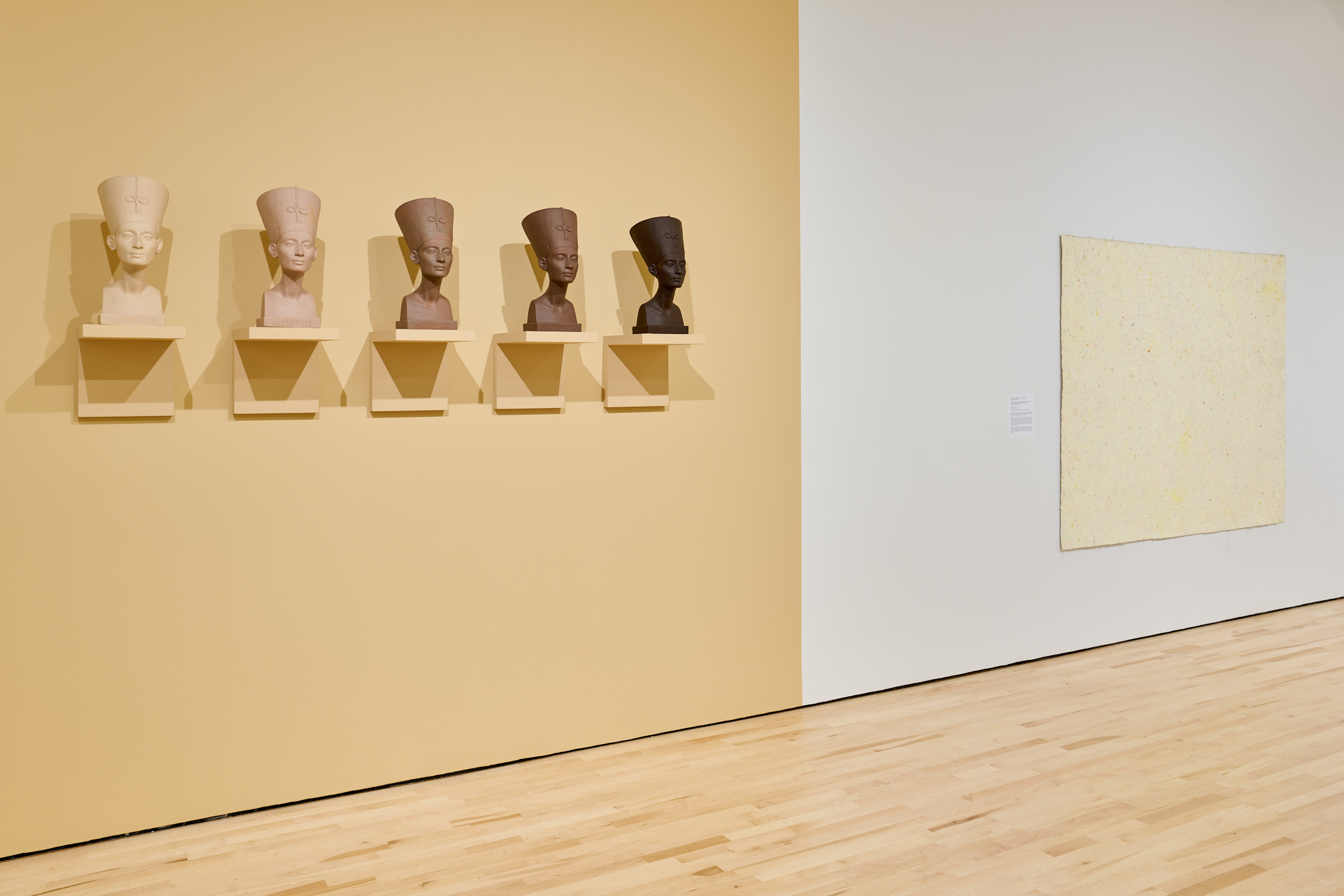 Beige wall with six Nefertiti statues in different skin tones on small shelves, light yellow abstract painting at right