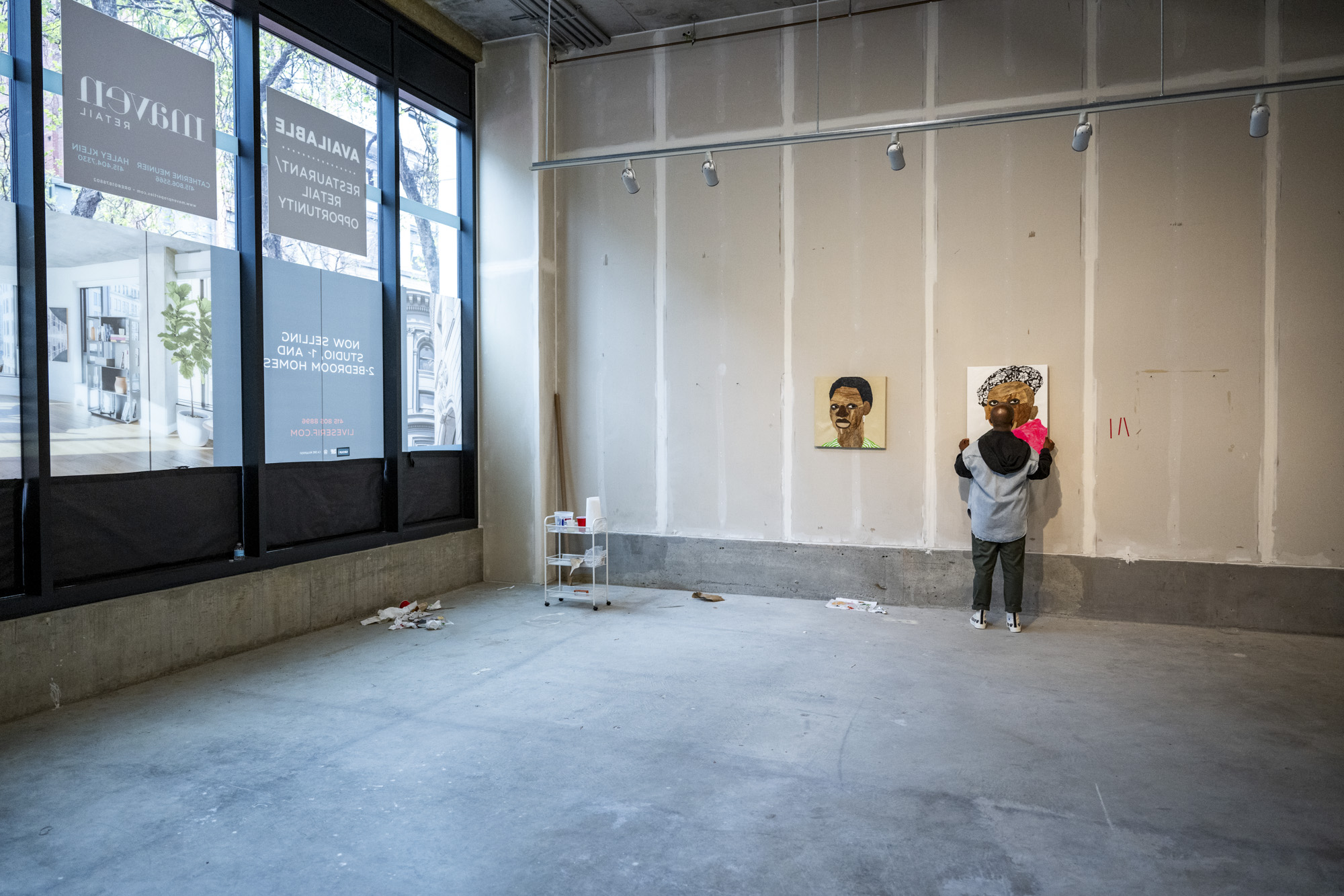 Person shifting an artwork on bare drywall in large street-facing space
