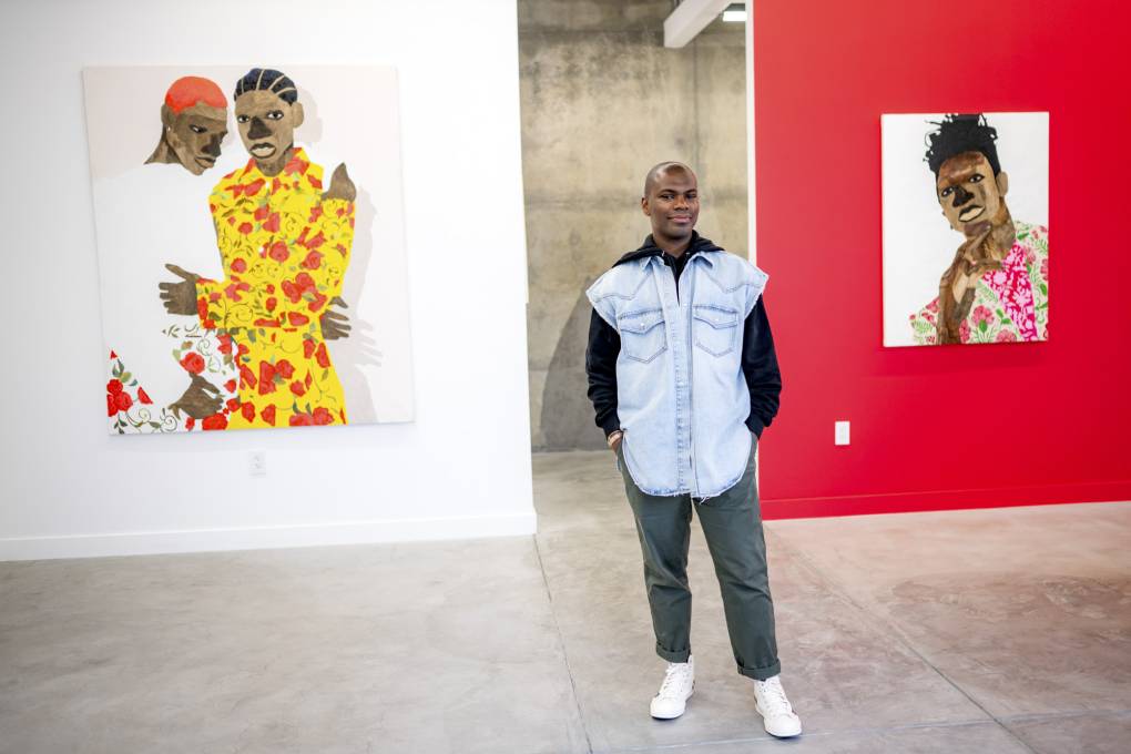 Black man stands with hands in pockets smiling in gallery space, two large paintings behind