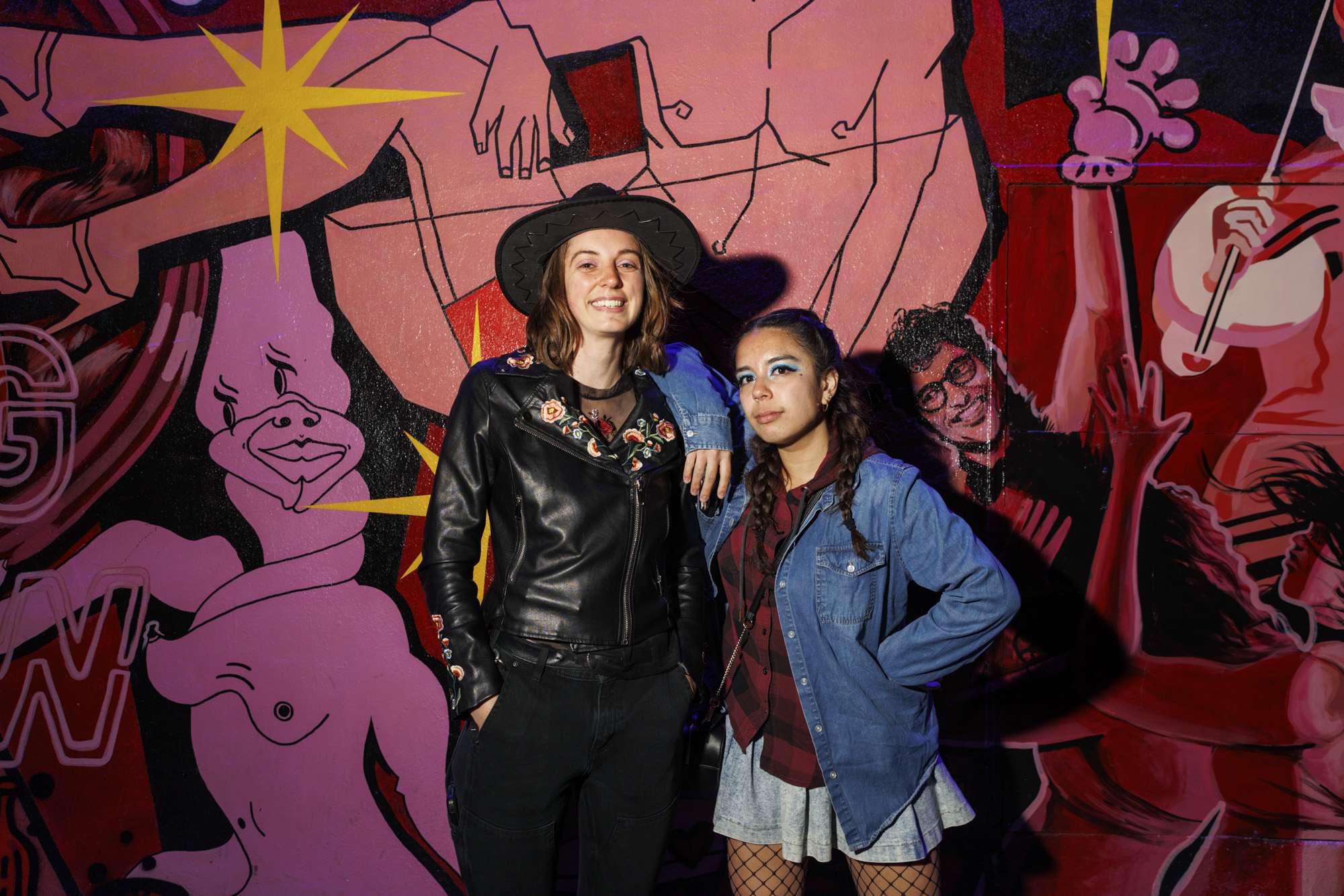 A smiling woman wearing a wide brimmed black hat and leather jacket stands with a shorter woman wearing her hair in braids and a short skirt. They are standing before a pink and red mural.