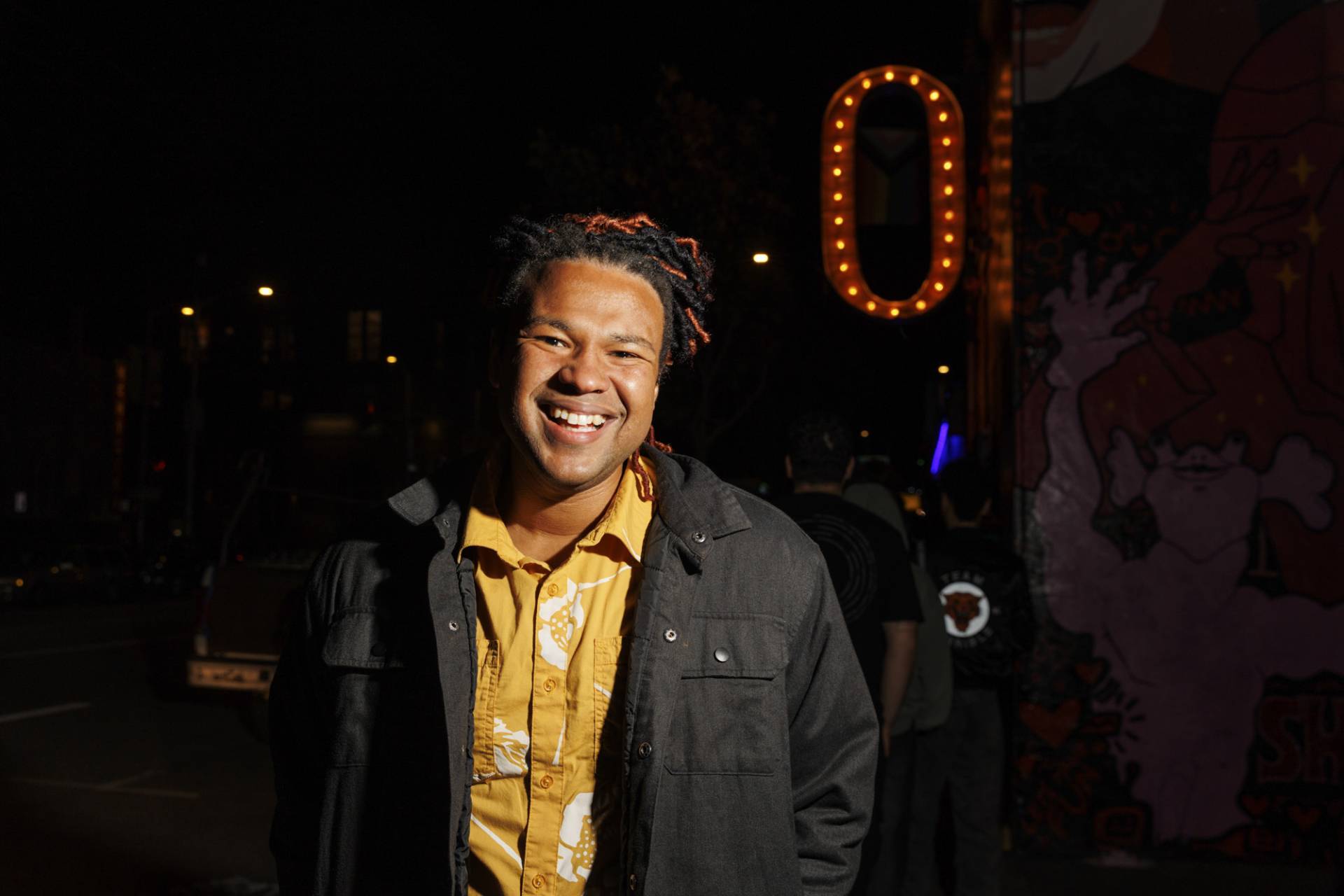 A Black man smiles broadly in front of a neon-lit O sign at night.