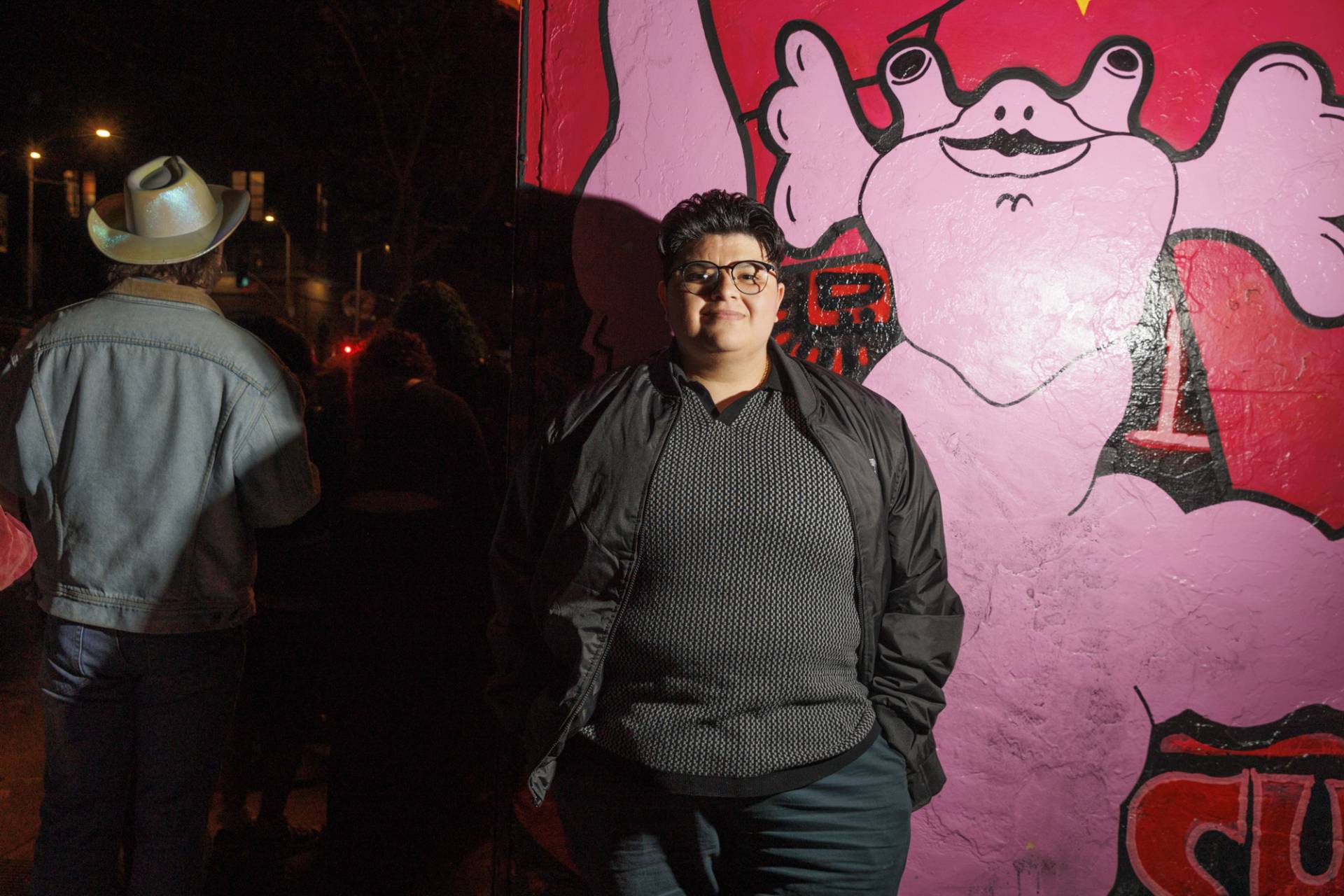 A person with glasses and short dark haircut, stands hands in pockets in front of a pink and red mural. 