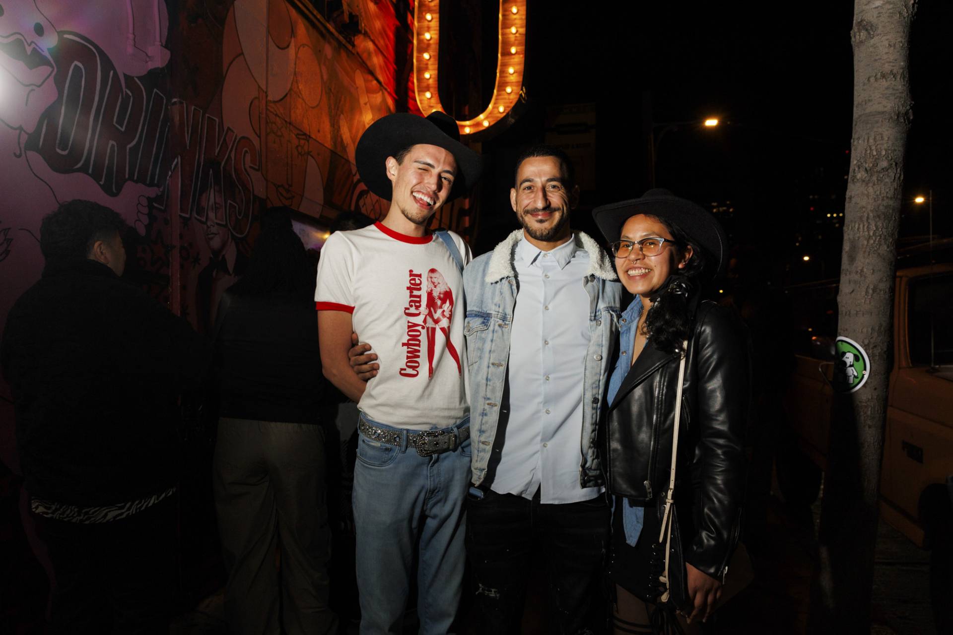 A slender white man in a Beyonce t-shirt, an Arabic man wearing a blue shirt and an Asian woman in a black leather jacket stand with arms around each other outside a nightclub.