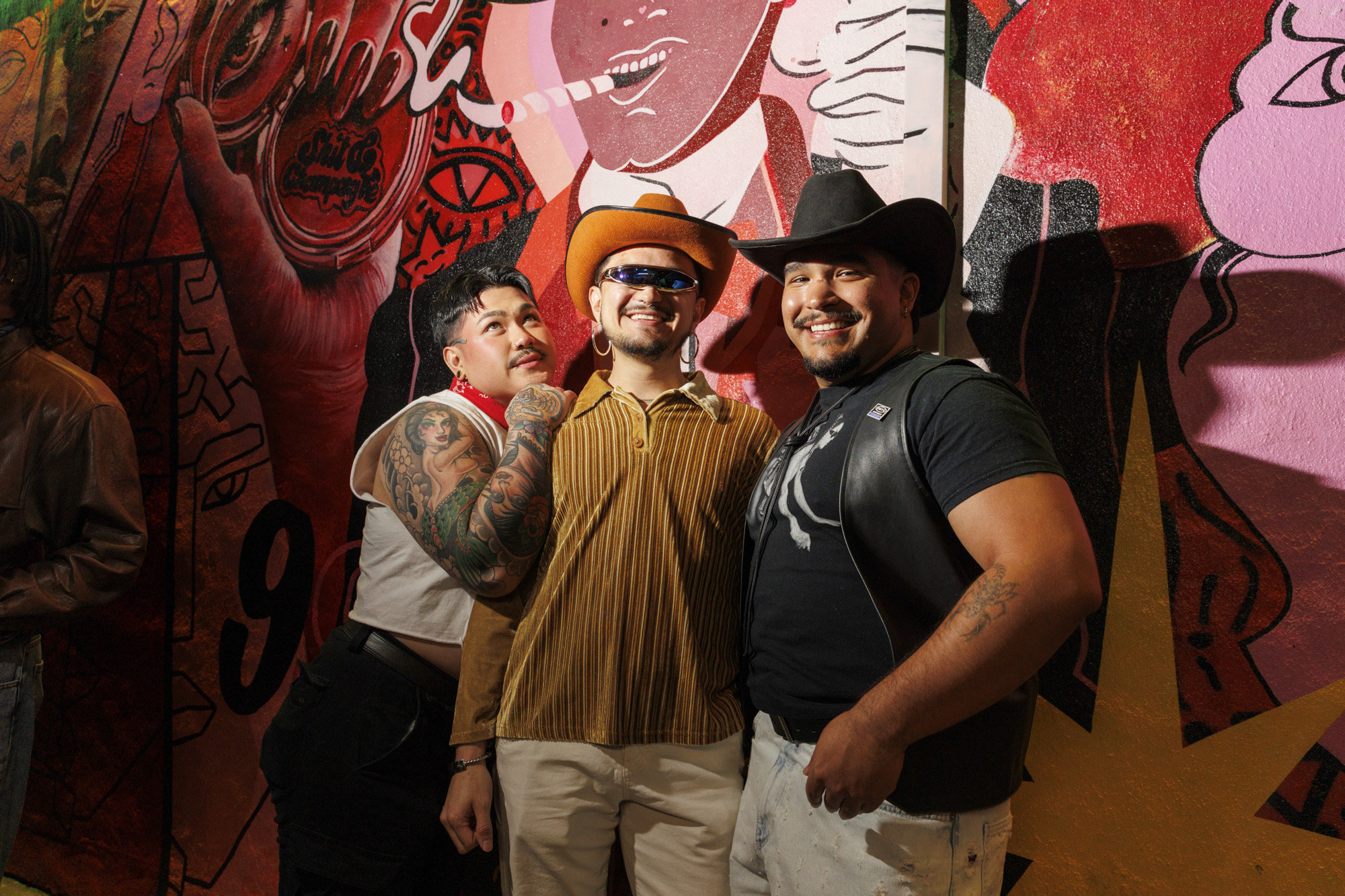 A trio of tattooed and flamboyant people, including two in cowboy hats, smile and line up before a pink and red mural.
