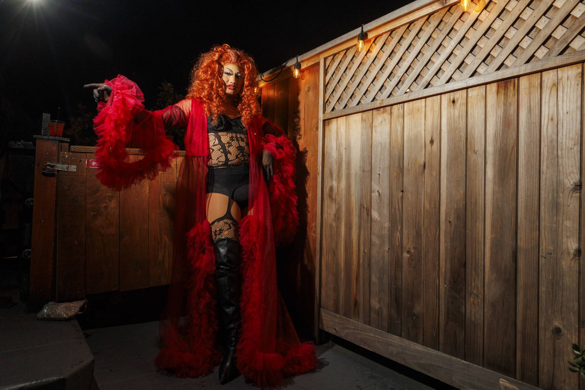 A drag queen strikes a pose in black lingerie and red dressing gown, next to a wooden fence decorated with lights.