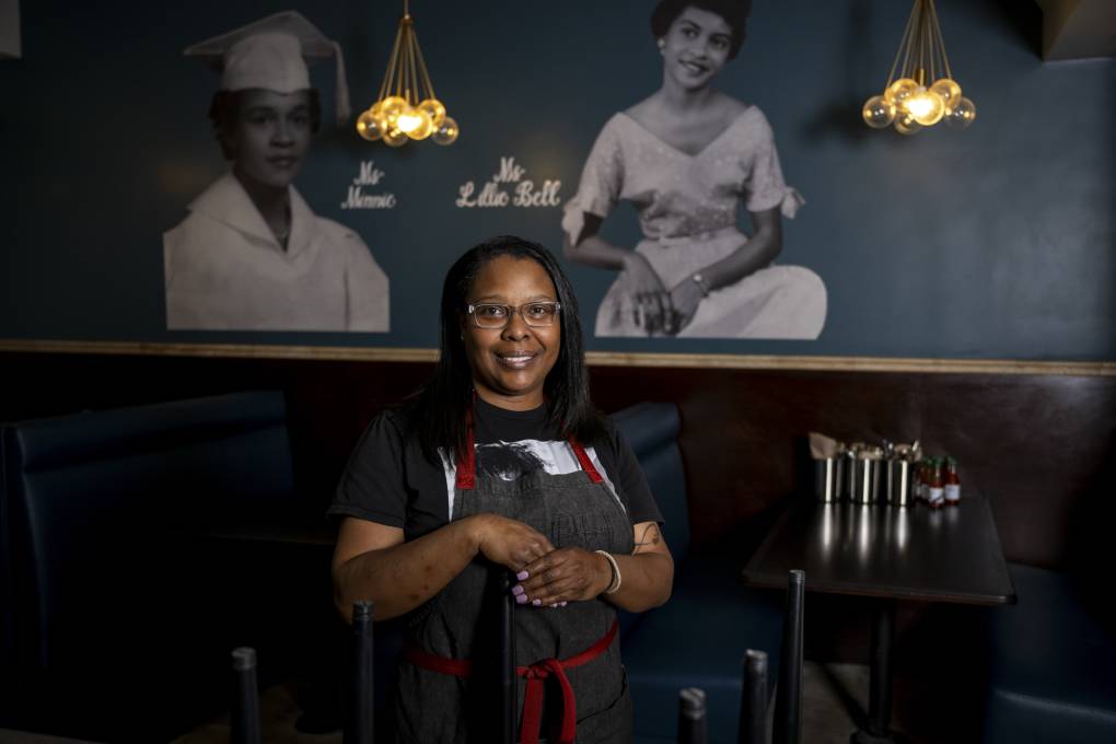 A chef poses inside her restaurant in front of a photo mural that shows her great-aunt and grandmother.
