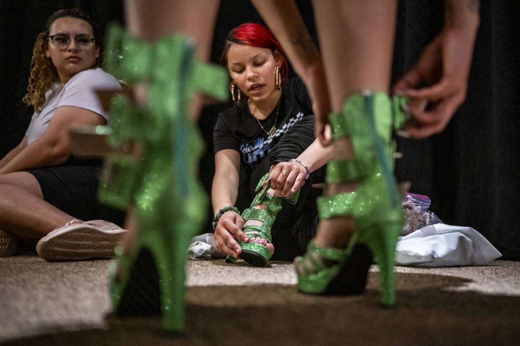 A young woman in black T-shirt and red dyed hair works on a costume, framed by two legs in green shoes.