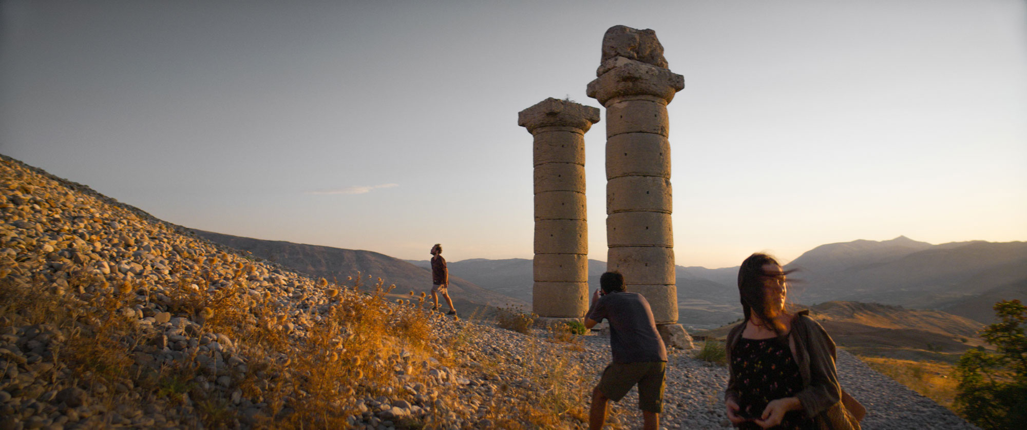 Three people in golden-lit landscape with ruins of two large columns
