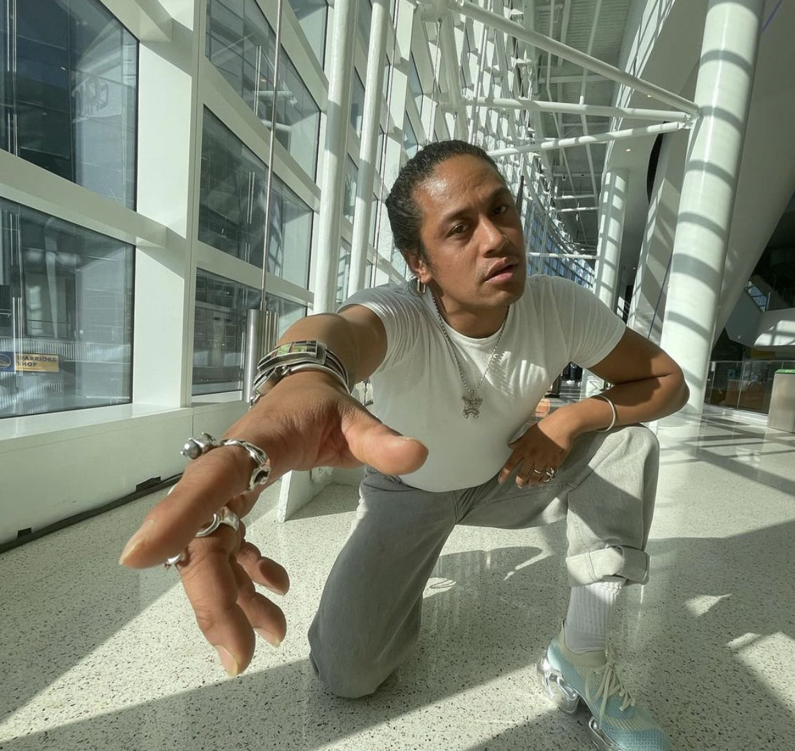 A person poses on one knee, looking directly at the camera with their right arm stretch forward toward the photographer.